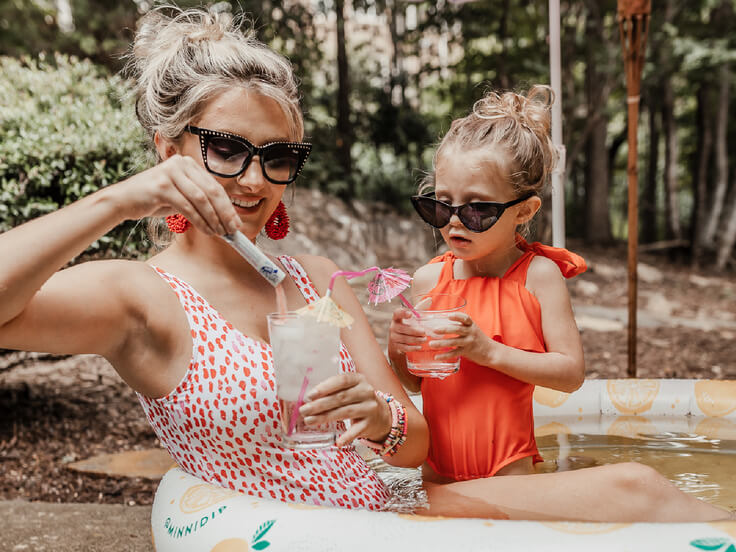 Flavored Water Drinks with Mom in the Kiddie Pool, A joyful outdoor scene with a woman and a young girl in bright, summer swimwear, wearing sunglasses, making drinks. The woman is stirring a glass with a raspberry lemonade drink mix, and the girl is holding a glass with a drink and a pink umbrella, both enjoying a sunny day by an inflatable pool surrounded by greenery