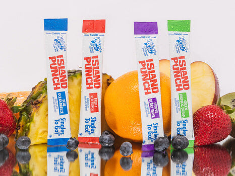 An assortment of Wyler’s Light Island Punch drink mix sticks standing upright, flanked by fresh fruits including pineapple, orange, apple, strawberries, and blueberries on a reflective surface, highlighting a colorful and healthy lifestyle choice