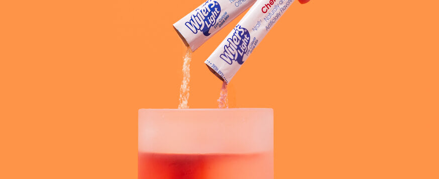 Mixing flavored water packets in a glass, Flavored water mixes, Two powdered drink packets pouring into a glass on an orange background