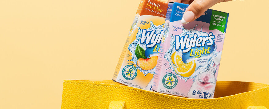 Hand holding Wyler's Light drink packets, Peach Iced Tea and Pink Lemonade flavors, inside a yellow tote bag, yellow background,  travel  sized water flavor packets